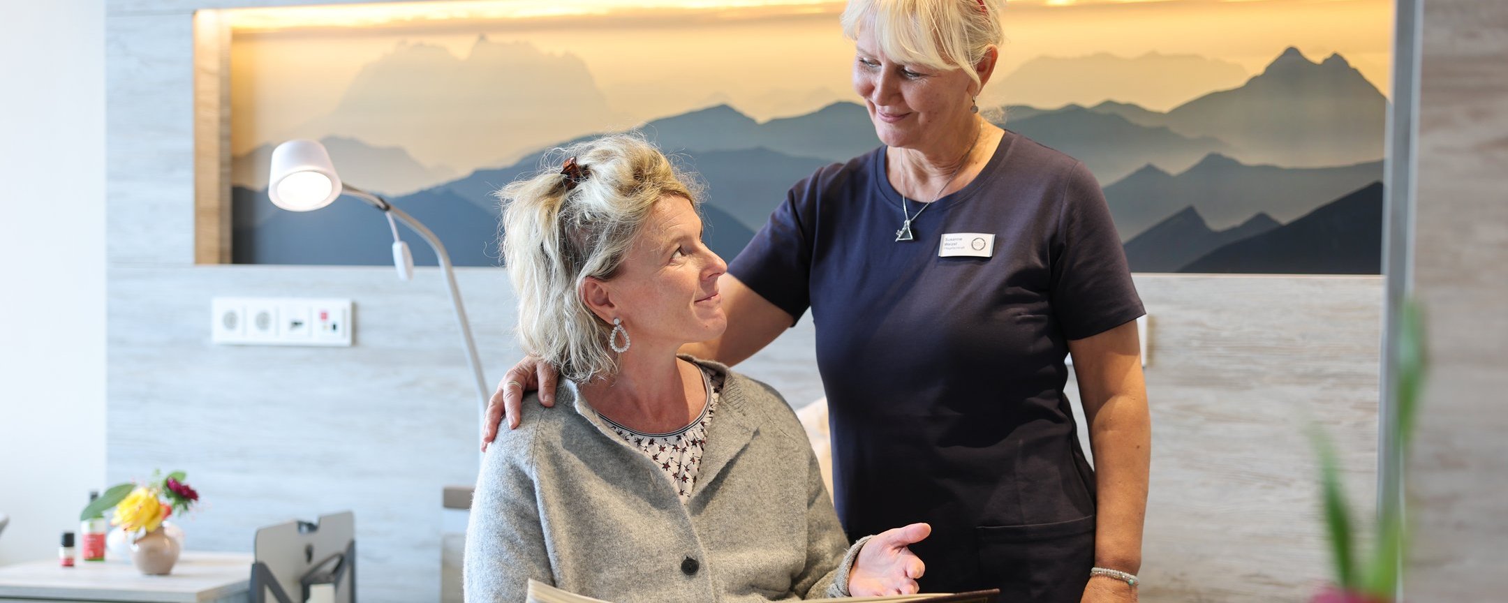 Eine Pflegekraft steht neben einer sitzenden Person im Gästezimmer des Oberland Hospiz, legt ihr beruhigend die Hand auf die Schulter.