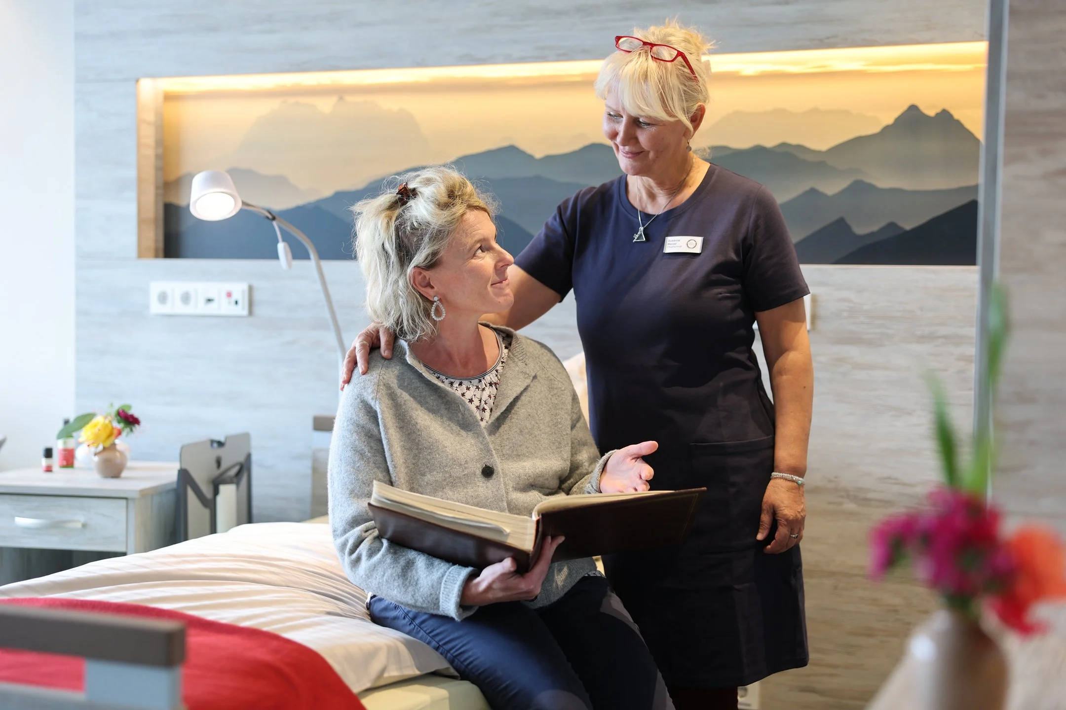 Eine Pflegekraft steht neben einer sitzenden Person im Gästezimmer des Oberland Hospiz, legt ihr beruhigend die Hand auf die Schulter.