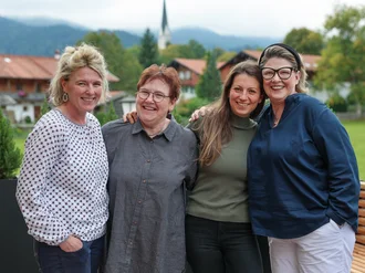 Vier Personen stehen gemeinsam lächelnd im Freien auf einer Terrasse mit Blick auf grüne Landschaft und Berge im Hintergrund.