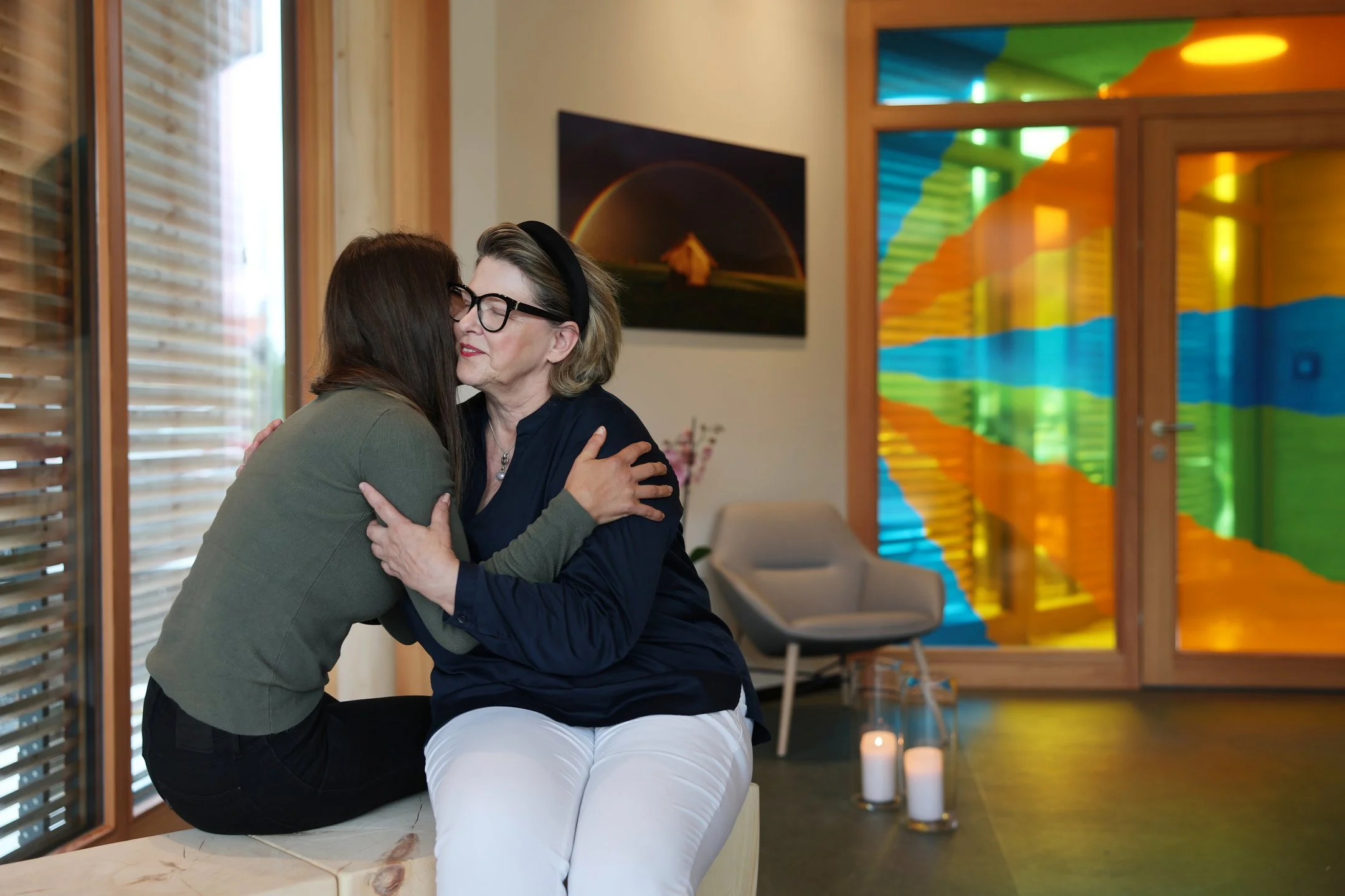 Zwei Menschen sitzen sich im Raum der Stille gegenüber und umarmen sich. Im Hintergrund buntes Glasfenster und warme, ruhige Atmosphäre.