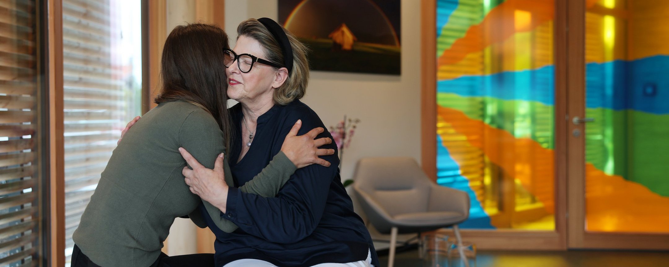 Zwei Menschen sitzen sich im Raum der Stille gegenüber und umarmen sich. Im Hintergrund buntes Glasfenster und warme, ruhige Atmosphäre.