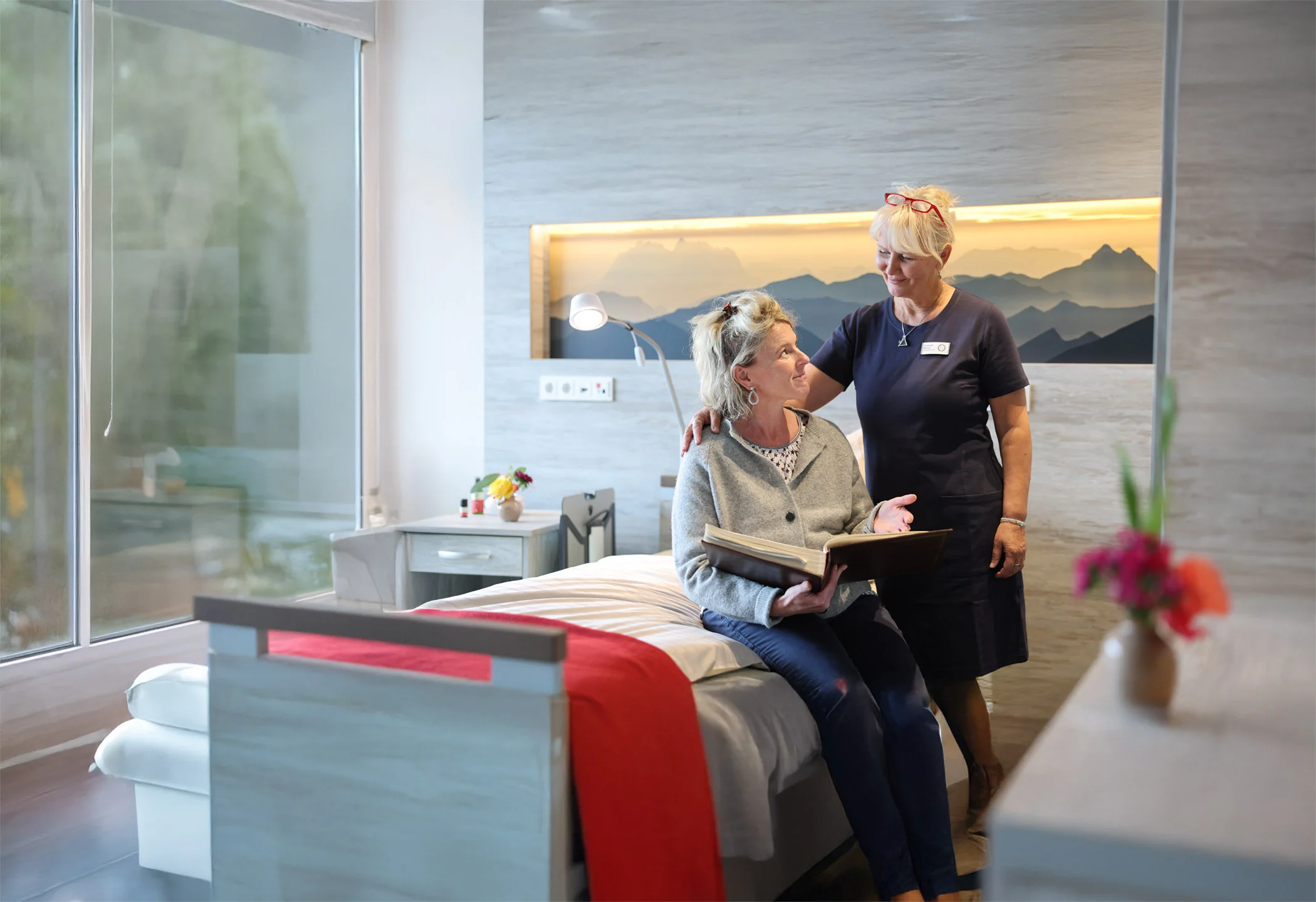 Eine Pflegekraft spricht lächelnd mit einer sitzenden Person im hellen Gästezimmer des Oberland Hospiz. Durch das große Fenster fällt Tageslicht.