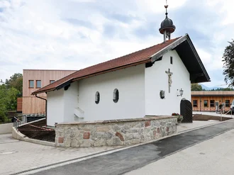Die kleine Kapelle vor dem Oberland Hospiz mit weißer Fassade, rotem Ziegeldach und Holzkreuz, umgeben von Bäumen und Weg.