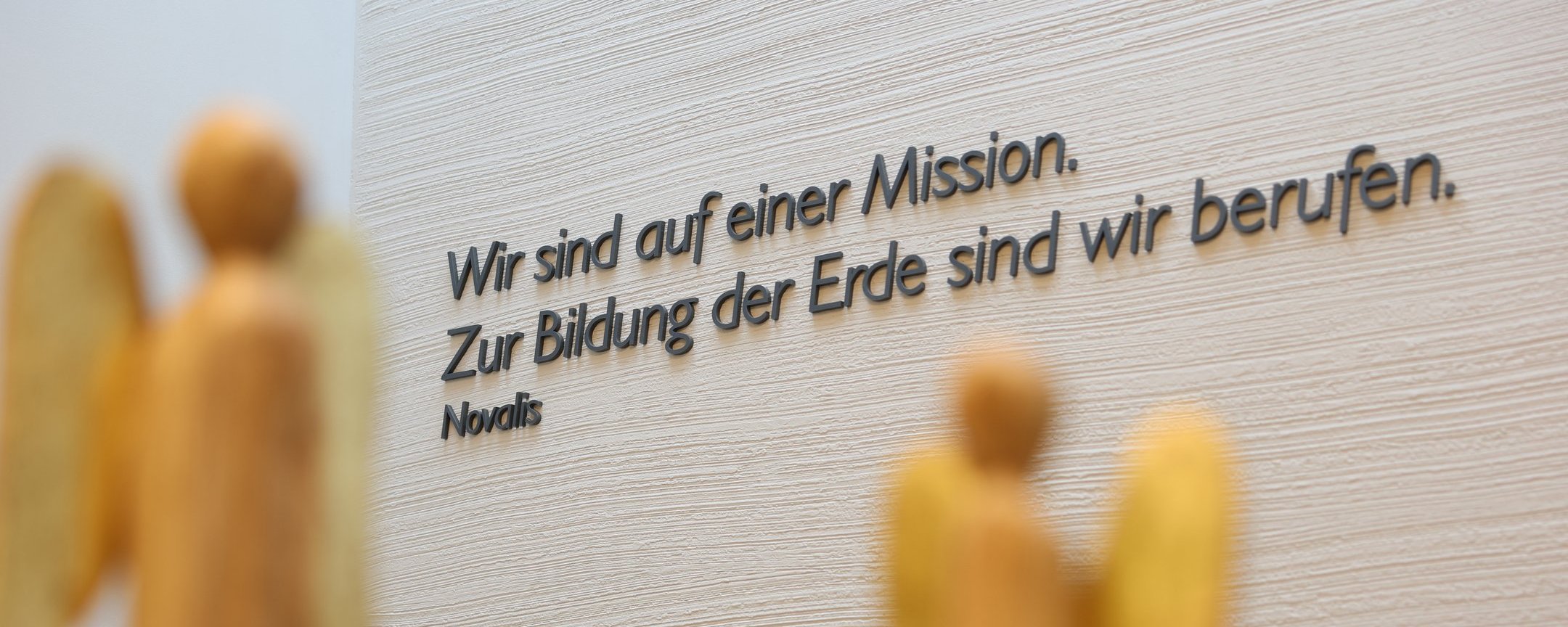 Zwei Holzfiguren in Engelgestalt stehen vor einer Wand mit dem Schriftzug „Wir sind auf einer Mission. Zur Erhaltung der Erde sind wir berufen."