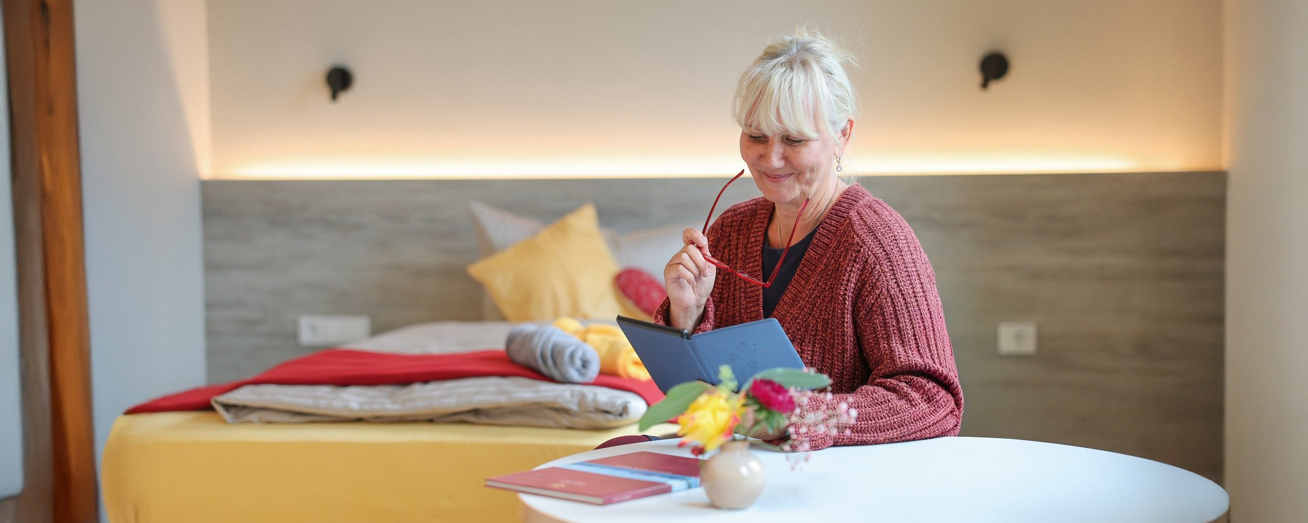 Eine Person sitzt in einem gemütlichen Gästezimmer des Oberland Hospiz an einem Tisch und bastelt oder schreibt. Auf dem Bett liegen farbige Kissen.