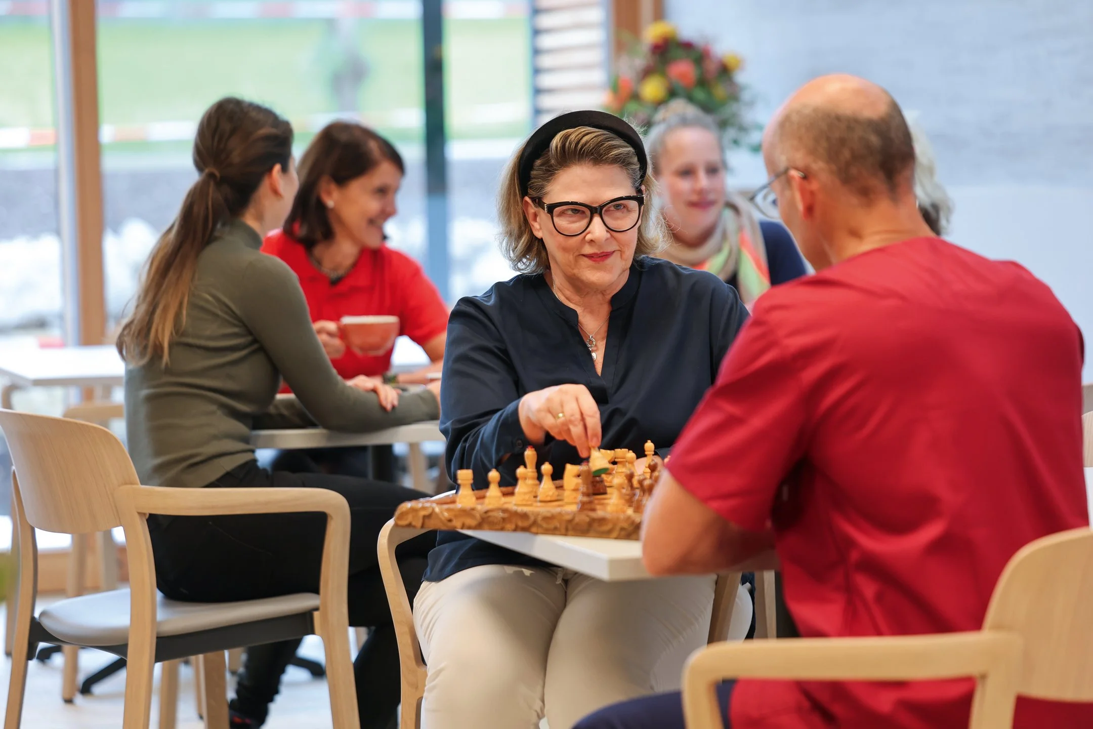 Zwei Personen spielen Schach an einem Tisch im hellen Gemeinschaftsraum des Oberland Hospiz. Im Hintergrund sitzen weitere Menschen bei Gesprächen.