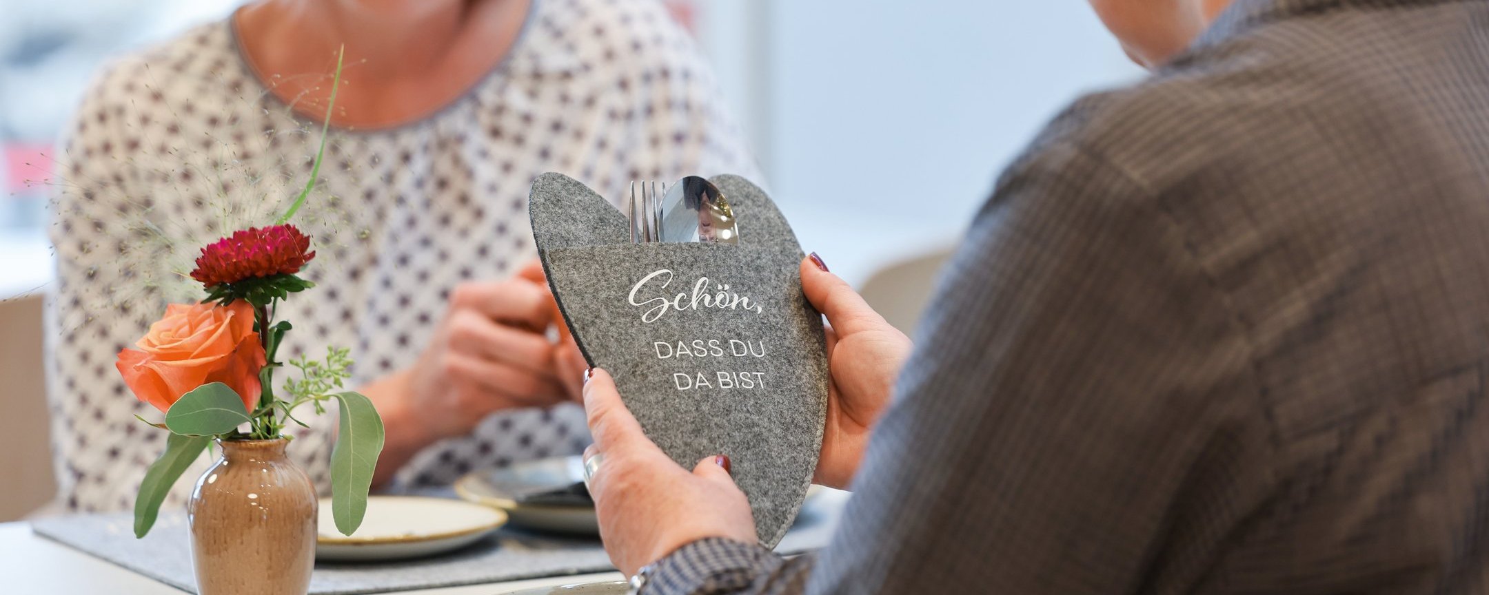 Zwei Personen sitzen an einem Tisch, eine hält eine herzförmige Steindekoration in den Händen, die andere lächelt. Auf dem Tisch steht eine kleine Blume.