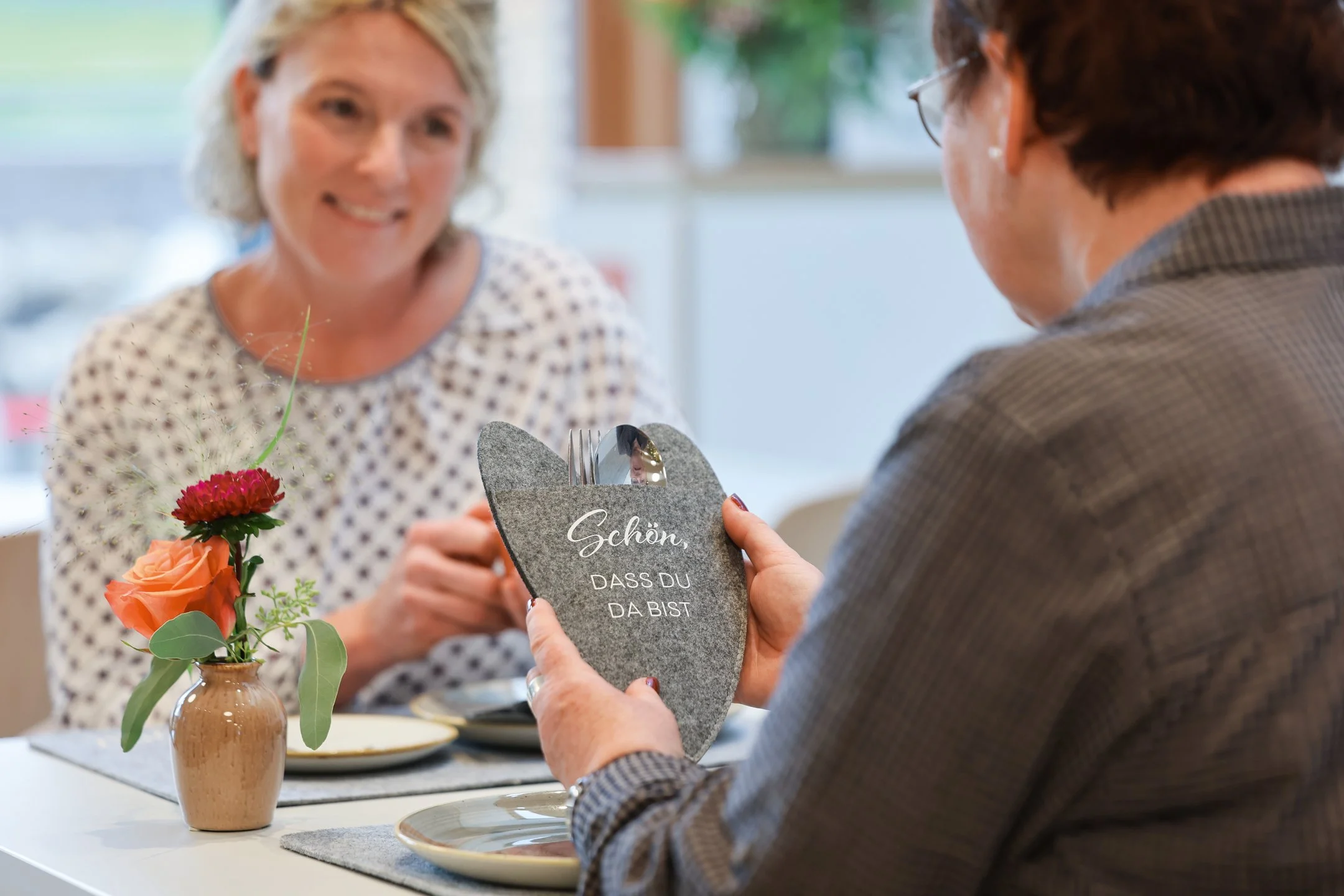 Zwei Personen sitzen an einem Tisch, eine hält eine herzförmige Steindekoration in den Händen, die andere lächelt. Auf dem Tisch steht eine kleine Blume.
