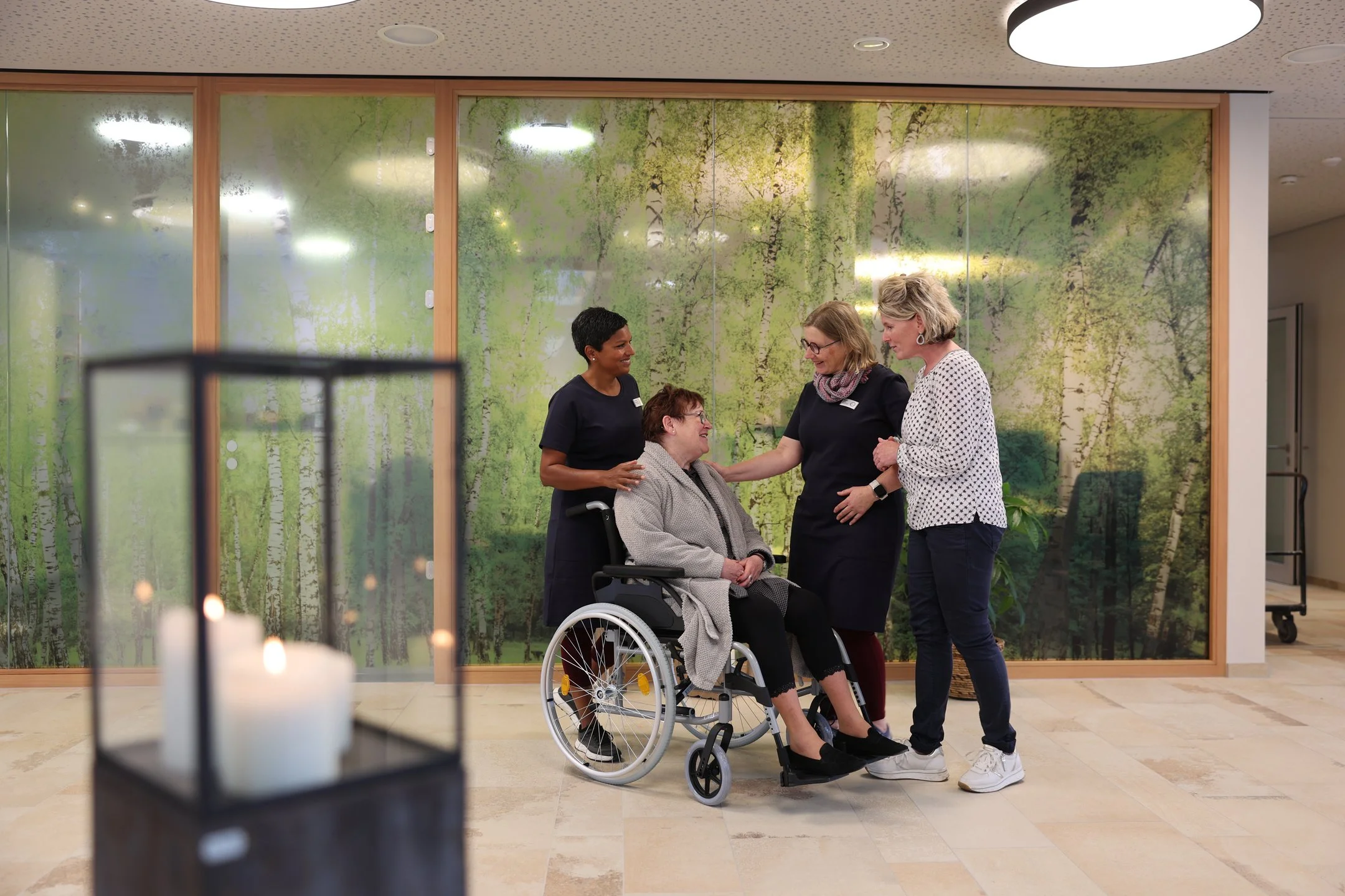 Vier Personen stehen und unterhalten sich mit einer Person im Rollstuhl im freundlich beleuchteten Aufenthaltsbereich des Oberland Hospiz.
