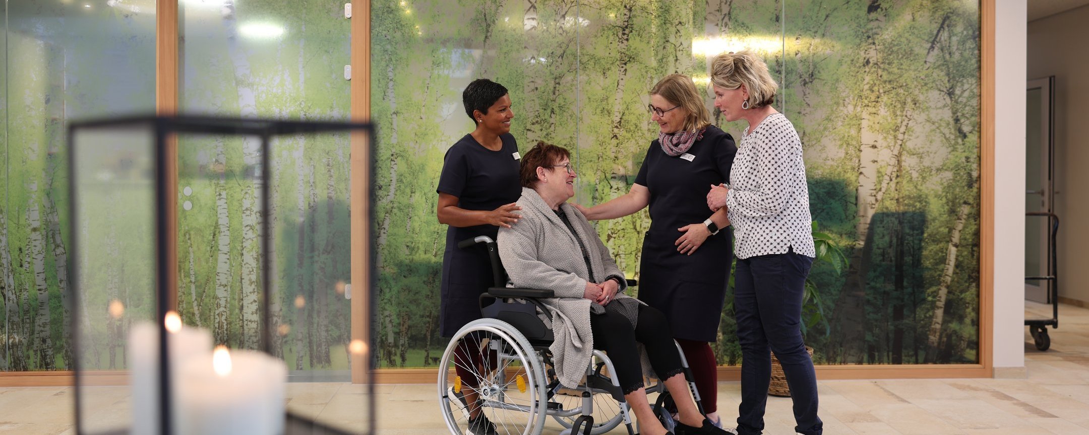Vier Personen stehen und unterhalten sich mit einer Person im Rollstuhl im freundlich beleuchteten Aufenthaltsbereich des Oberland Hospiz.