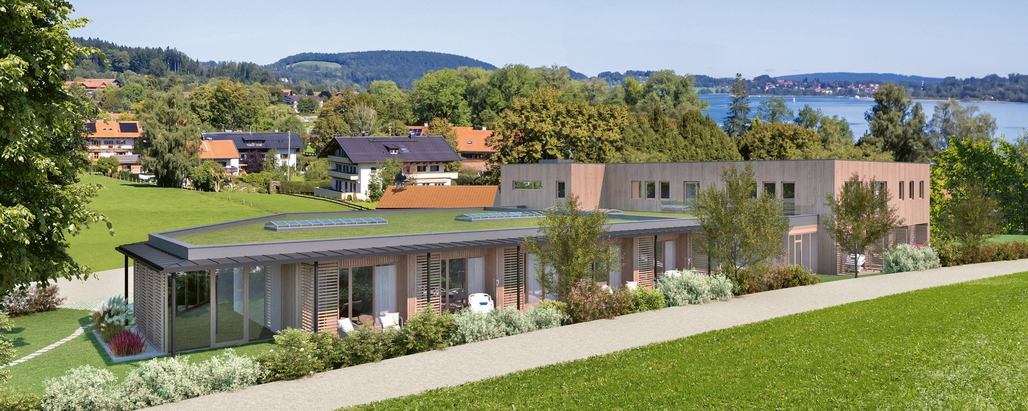 Das moderne Gebäude des Oberland Hospiz liegt auf einer grünen Anhöhe mit Blick auf Bad Wiessee und den Tegernsee, umgeben von Bäumen und Wiesen.
