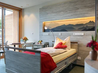 Helles, modern eingerichtetes Einzelzimmer im Oberland Hospiz mit Pflegebett, Blumen, Holzdetails und Blick auf eine Terrasse mit Sitzgelegenheit.