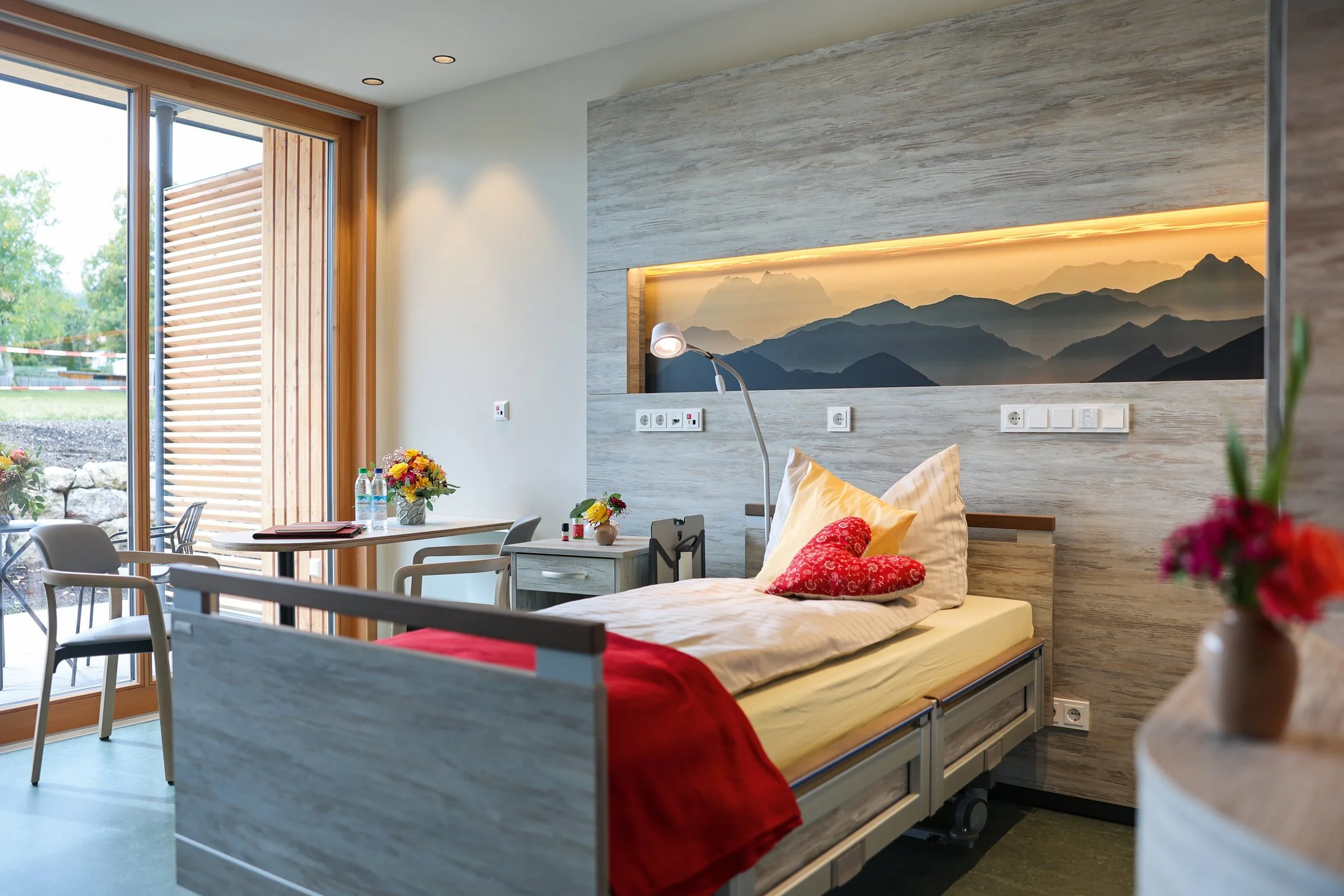 Helles, modern eingerichtetes Einzelzimmer im Oberland Hospiz mit Pflegebett, Blumen, Holzdetails und Blick auf eine Terrasse mit Sitzgelegenheit.
